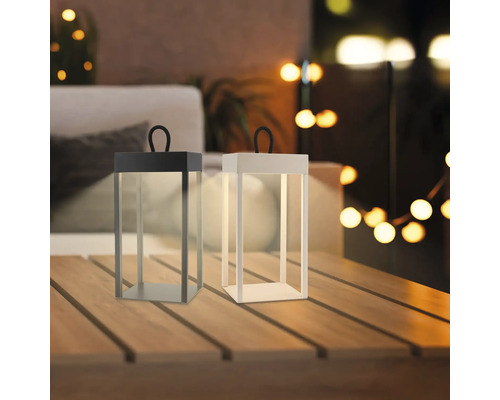 Zwei moderne LED Tischleuchten in Laternenform mit Tragegriff stehen auf einem Holztisch auf einer Terrasse in abendlicher Atmosphäre.