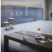 Moderne Tischleuchte auf einem Holztisch mit Kaffeetasse und Zeitschrift auf einer Terrasse mit Laternen im Hintergrund