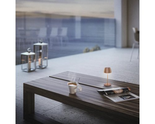 Moderne Tischleuchte auf einem Holztisch mit Kaffeetasse und Zeitschrift auf einer Terrasse mit Laternen im Hintergrund