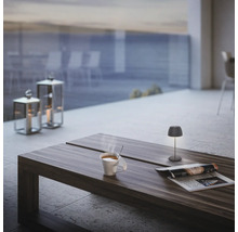 Moderne Tischleuchte auf Holztisch auf einer Terrasse mit dampfender Tasse und Zeitschrift, im Hintergrund Windlichter und Meerblick.