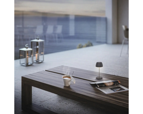 Moderne Tischleuchte auf Holztisch auf einer Terrasse mit dampfender Tasse und Zeitschrift, im Hintergrund Windlichter und Meerblick.