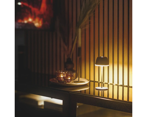 Eine kleine Tischlampe leuchtet neben Teelichtern auf einem Tablett vor einer Wand mit Holzlamellen.