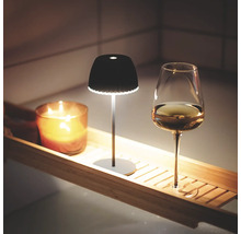 Eine kleine, moderne LED Akkuleuchte steht zusammen mit einer Kerze und einem Weinglas auf einer Holz Badewannenablage.