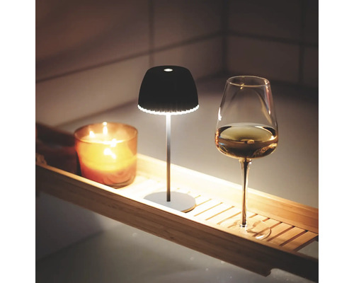 Eine kleine, moderne LED Akkuleuchte steht zusammen mit einer Kerze und einem Weinglas auf einer Holz Badewannenablage.