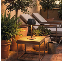 Moderne kabellose LED Tischleuchte auf einem kleinen Holztisch auf einer Terrasse mit Pflanzen und Gartenliegen bei Dämmerung.