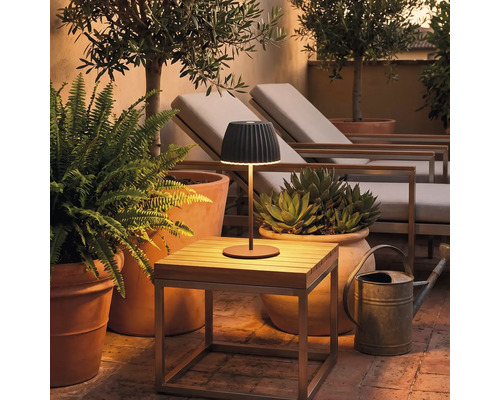 Moderne kabellose LED Tischleuchte auf einem kleinen Holztisch auf einer Terrasse mit Pflanzen und Gartenliegen bei Dämmerung.