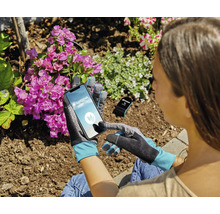 Une personne porte des gants de jardinage et utilise un smartphone avec l'application Gardena Smart System dans un jardin avec des fleurs.