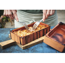 Une personne effiloche de la viande braisée avec deux fourchettes dans un plat en terre cuite rectangulaire sur une planche en bois.