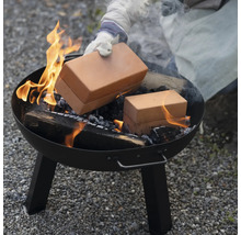 Une personne place une boîte de cuisson en brique dans un brasero rond avec du bois en feu.