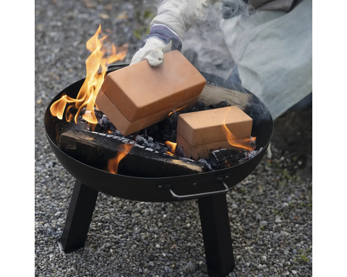 Une personne place une boîte de cuisson en brique dans un brasero rond avec du bois en feu.