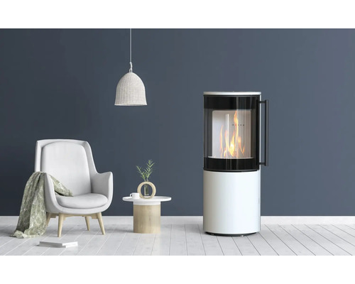 Poêle à bois moderne de forme cylindrique avec vitre et feu ardent dans un salon, à côté d'un fauteuil et d'une table d'appoint.