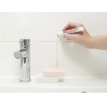 Une main utilise un marqueur pour joints Edding afin de rafraîchir les joints de carrelage sur un mur près d'un robinet.