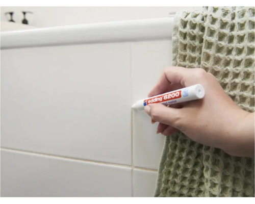 Personne utilisant un feutre à joints Edding 8200 pour rafraîchir les joints de carrelage sur un mur de salle de bain.