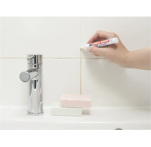 Une main utilise un marqueur de joints Edding pour rafraîchir les joints de carrelage sur un mur au-dessus d'un lavabo avec robinetterie.