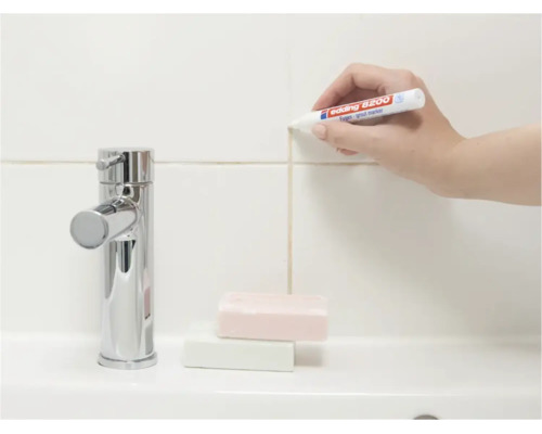 Une main utilise un marqueur de joints Edding pour rafraîchir les joints de carrelage sur un mur au-dessus d'un lavabo avec robinetterie.