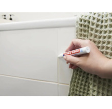 Une main utilise un marqueur pour joints Edding 8200 pour retoucher les joints de carrelage sur un mur de salle de bain.