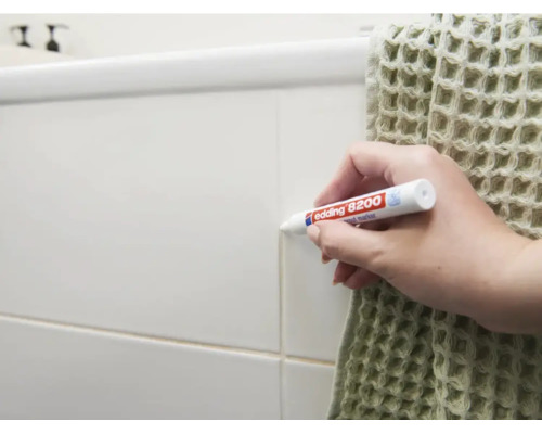 Une main utilise un marqueur pour joints Edding 8200 pour retoucher les joints de carrelage sur un mur de salle de bain.
