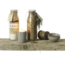 Deux bouteilles en verre avec des préparations pour gâteaux, une bougie dans un pot, une bobine de fil et des petits bols sur une planche en bois.