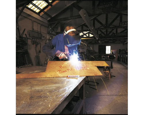 Un soudeur travaille sur une plaque métallique avec un marqueur Edding dans un atelier industriel.