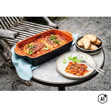 Eine rechteckige Keramik Auflaufform mit frisch zubereiteter Lasagne auf einem runden Tisch mit Teller, Brot und Besteck. (Image AI modified or generated)