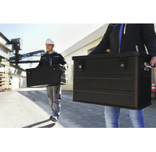 Zwei Arbeiter tragen schwarze Aluminiumboxen von Alutec vor einem Industriegelände.
