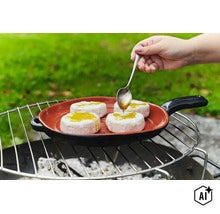 Eine Hand giesst Öl mit einem Löffel über Camembert Käse in einer gusseisernen Grillpfanne auf einem Holzkohlegrill im Freien. (Image AI modified or generated)