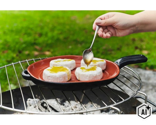 Eine Hand giesst Öl mit einem Löffel über Camembert Käse in einer gusseisernen Grillpfanne auf einem Holzkohlegrill im Freien. (Image AI modified or generated)