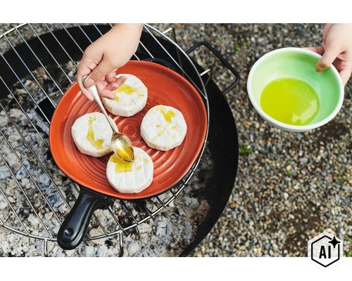 Person beträufelt auf einem Grill stehenden Grillkäse in einer Keramikpfanne mit Öl. (Image AI modified or generated)