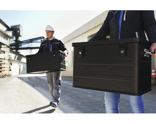 Zwei Arbeiter tragen schwarze Aluminiumboxen von Alutec vor einem Industriegelände.
