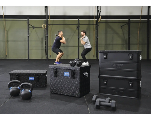 Drei schwarze Aluminiumboxen von Alutec Munchen auf dem Boden eines Fitnessstudios mit Sportgeräten wie Kurzhanteln und zwei trainierenden Männern.