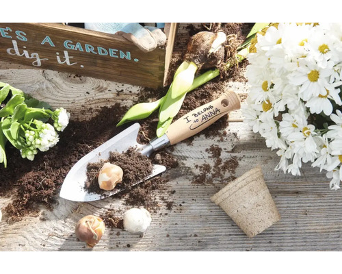 Personalisierte Handschaufel aus Metall mit Holzgriff und Blumenzwiebeln auf einem Holztisch mit Pflanzenarrangement.