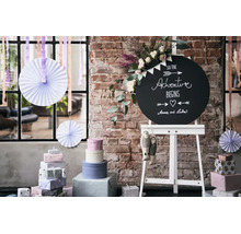 Un tableau noir rond avec inscription sur un chevalet, entouré de piles de cadeaux, d’éventails en papier et de fleurs devant un mur en briques.