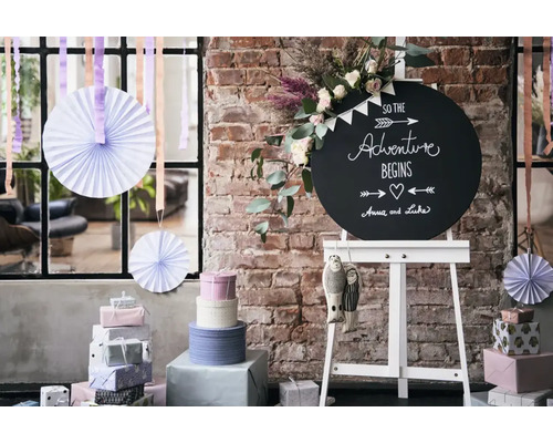 Un tableau noir rond avec inscription sur un chevalet, entouré de piles de cadeaux, d’éventails en papier et de fleurs devant un mur en briques.