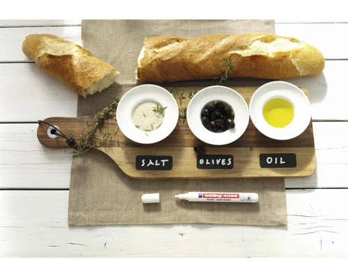 Un marqueur craie edding posé sur une planche en bois avec des bols étiquetés pour le sel, les olives et l'huile à côté d'une baguette.