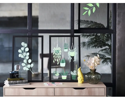 Deux cadres en verre noirs décorés de dessins de plantes, posés sur une commode claire avec des objets décoratifs devant une paroi vitrée.