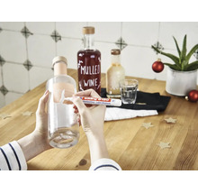 Une main utilise un stylo effaceur d etiquettes Edding 8180 pour enlever une étiquette d une bouteille en verre sur une table en bois.