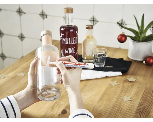 Une main utilise un stylo effaceur d etiquettes Edding 8180 pour enlever une étiquette d une bouteille en verre sur une table en bois.