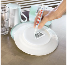 Une personne utilise un stylo dissolvant d etiquette Edding 8180 pour retirer une etiquette code barre sous une assiette sur une table en bois.