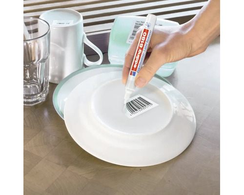 Une personne utilise un stylo dissolvant d etiquette Edding 8180 pour retirer une etiquette code barre sous une assiette sur une table en bois.