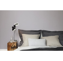 Modernes Schlafzimmer mit Wand in neutralem Grau, Holznachtisch, Wandleuchte und Bett mit verschiedenfarbigen Textilien.
