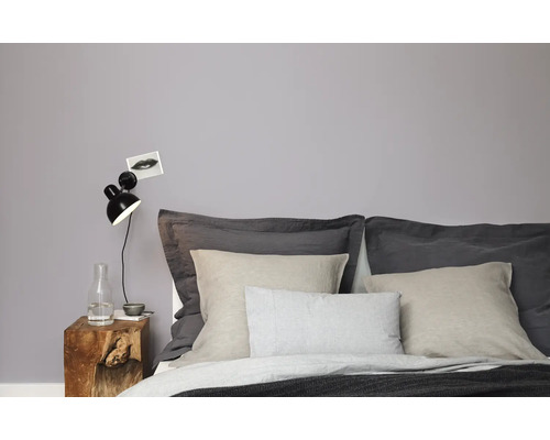 Modernes Schlafzimmer mit Wand in neutralem Grau, Holznachtisch, Wandleuchte und Bett mit verschiedenfarbigen Textilien.