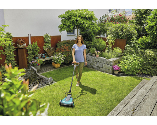 Eine Frau benutzt einen Rasenmäher in einem gepflegten Garten mit Steingarten und üppiger Bepflanzung.