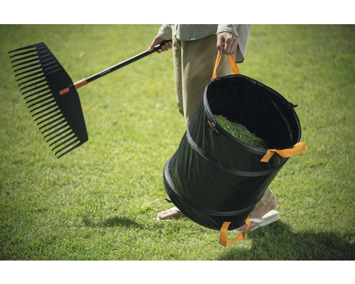 Une personne porte un râteau et un sac de jardin sur une pelouse.