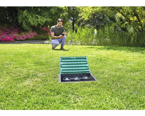 Paysage de jardin avec un homme utilisant un smartphone, au premier plan un boîtier de distribution électrique encastré dans la pelouse avec un couvercle ouvert.