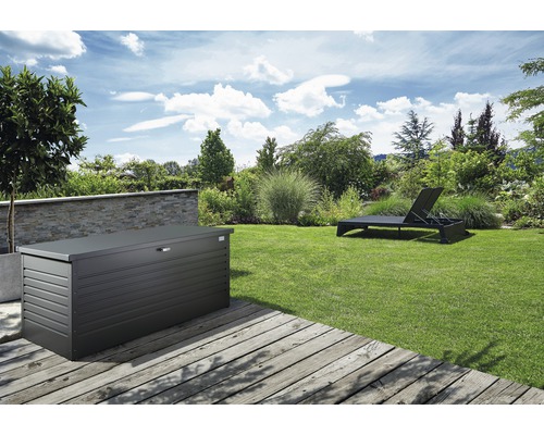 Scène de jardin avec un coffre de rangement en métal sur une terrasse en bois et une chaise longue sur la pelouse