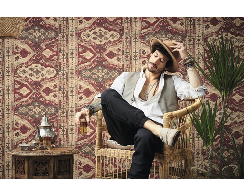 Mann sitzt in einem Korbsessel vor einer Wand mit einem roten Muster.