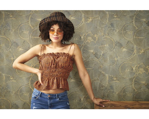 Femme portant un chapeau, un haut à motifs et un jean devant un papier peint à motifs