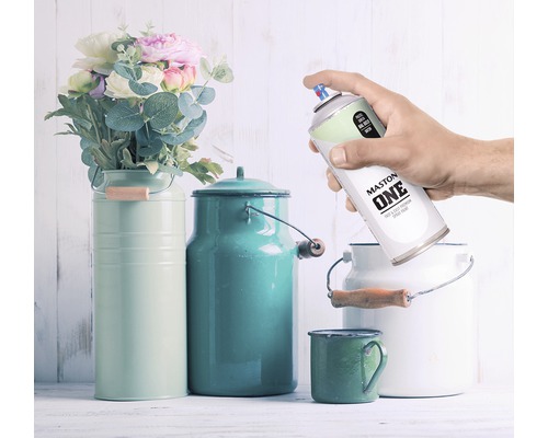 Scène décorative avec des fleurs, des bidons à lait colorés et une main tenant un aérosol Maston One.