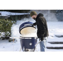 Un homme ouvre un barbecue en céramique bleu dans un jardin enneigé