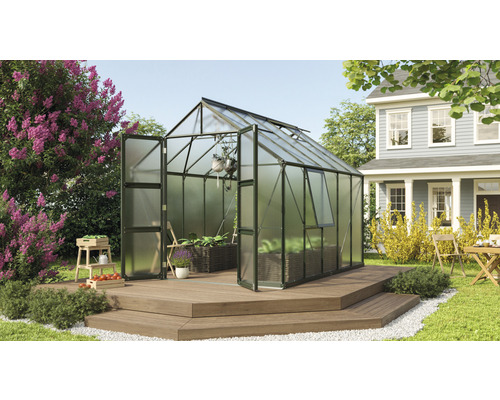 Gewächshaus Vitavia Olymp 8300 Hohlkammerplatten 4 mm 257 x 324 cm smaragd Gewächshaus mit geöffneten Türen und Pflanzen auf einer Holzterrasse in einem Garten.